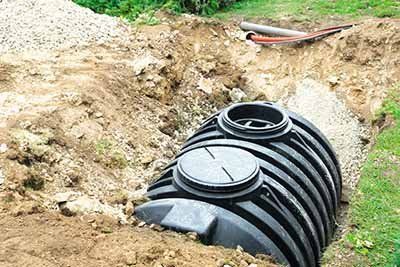 A black plastic septic tank is partially buried in a dirt trench, showing two access ports on top.