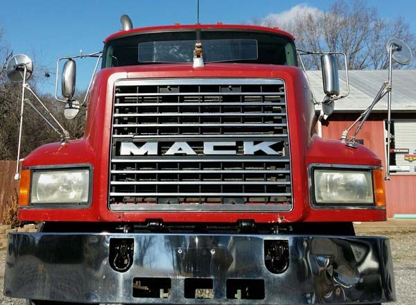 Front view of a red Mack semi-truck cab with a chrome grille and bumper in a rural outdoor setting.