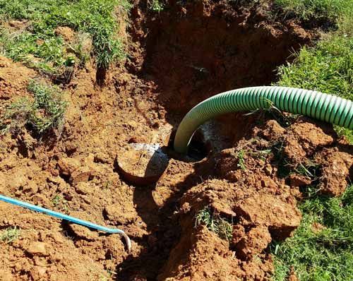 A green suction hose inserted into an open, dirt-excavated hole above a buried septic tank access lid.
