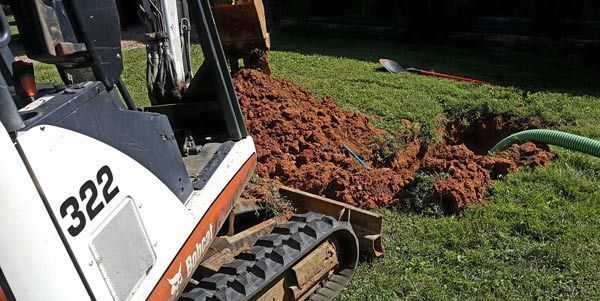 A Bobcat 322 excavator sits next to a hole dug in a grassy yard, with a green hose extending into the pit.