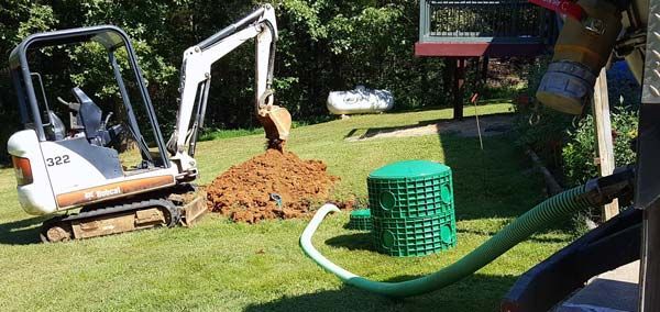 A small white excavator digs in a grassy yard near a green access riser and a drainage hose.