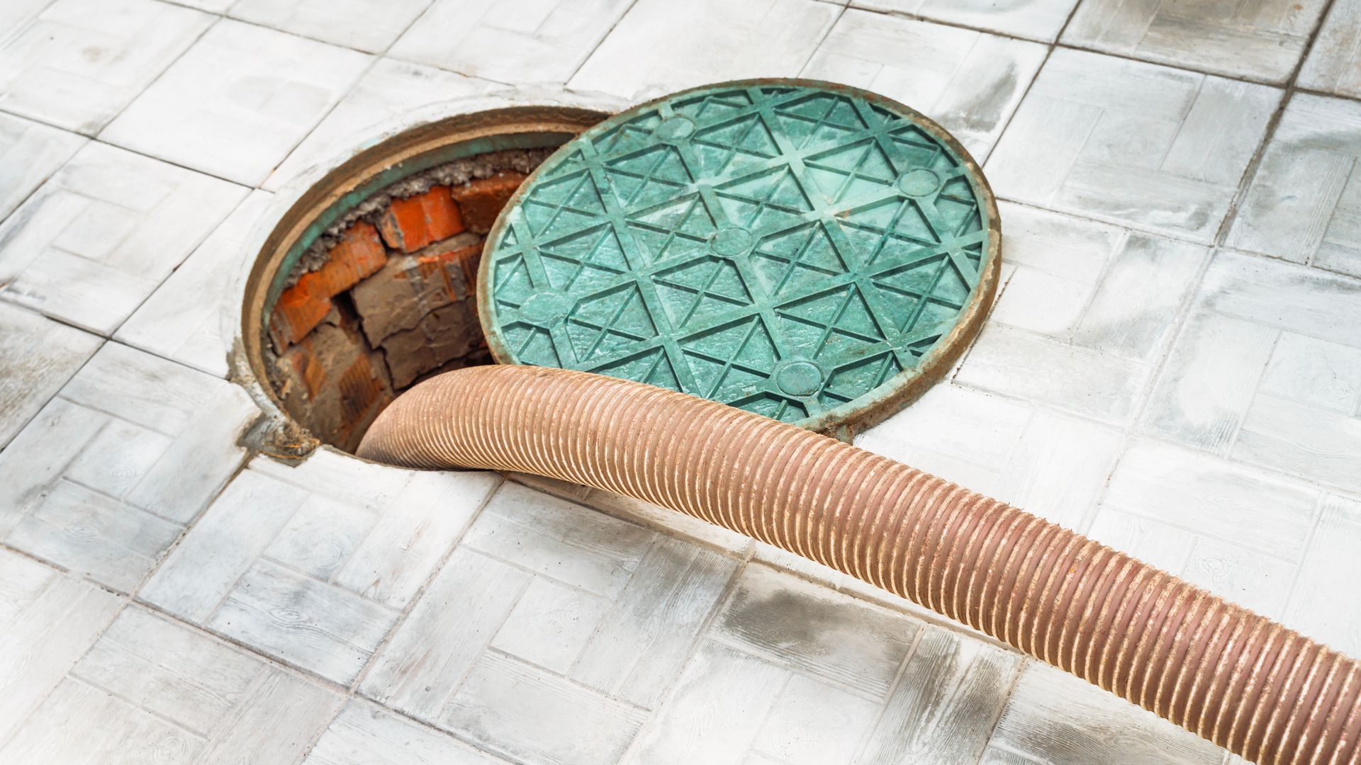 A suction hose extends into an open maintenance manhole with a green circular cover, set in a light-colored paved area.