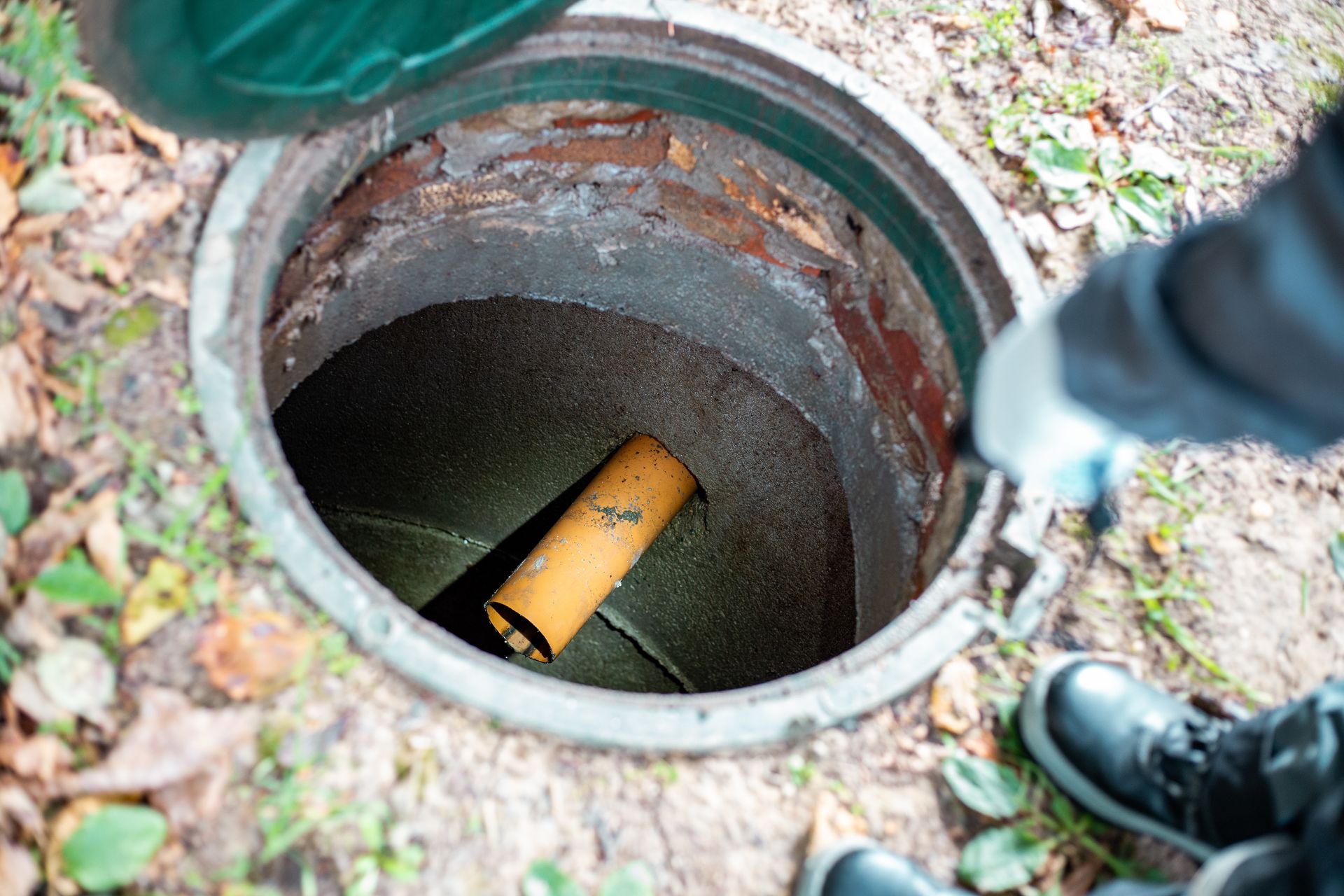 A person wearing gloves looks into an open, round concrete utility access shaft containing a yellow pipe.