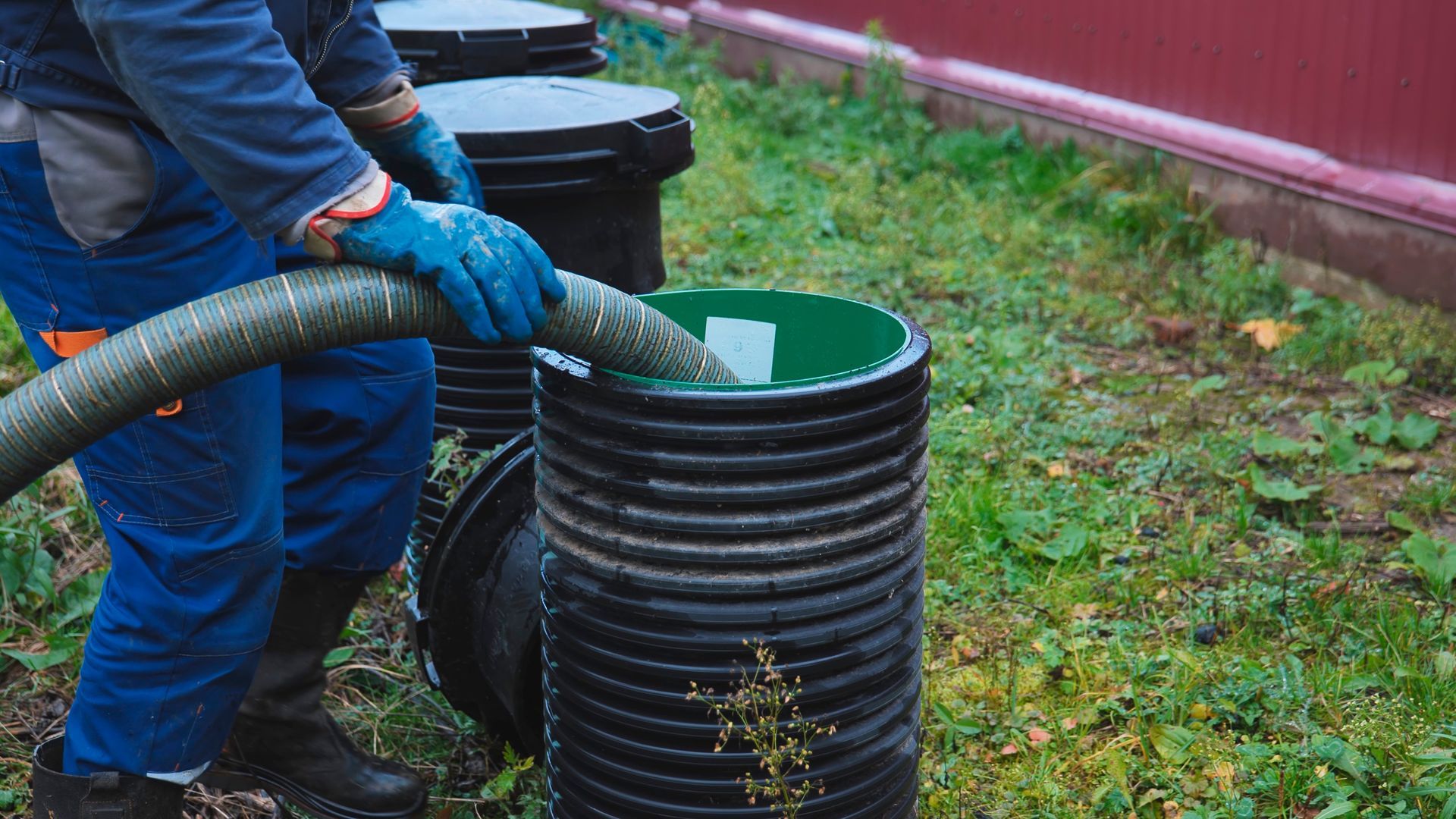 A worker in blue workwear and gloves uses a hose to pump liquid from a black corrugated septic riser in a grassy yard.