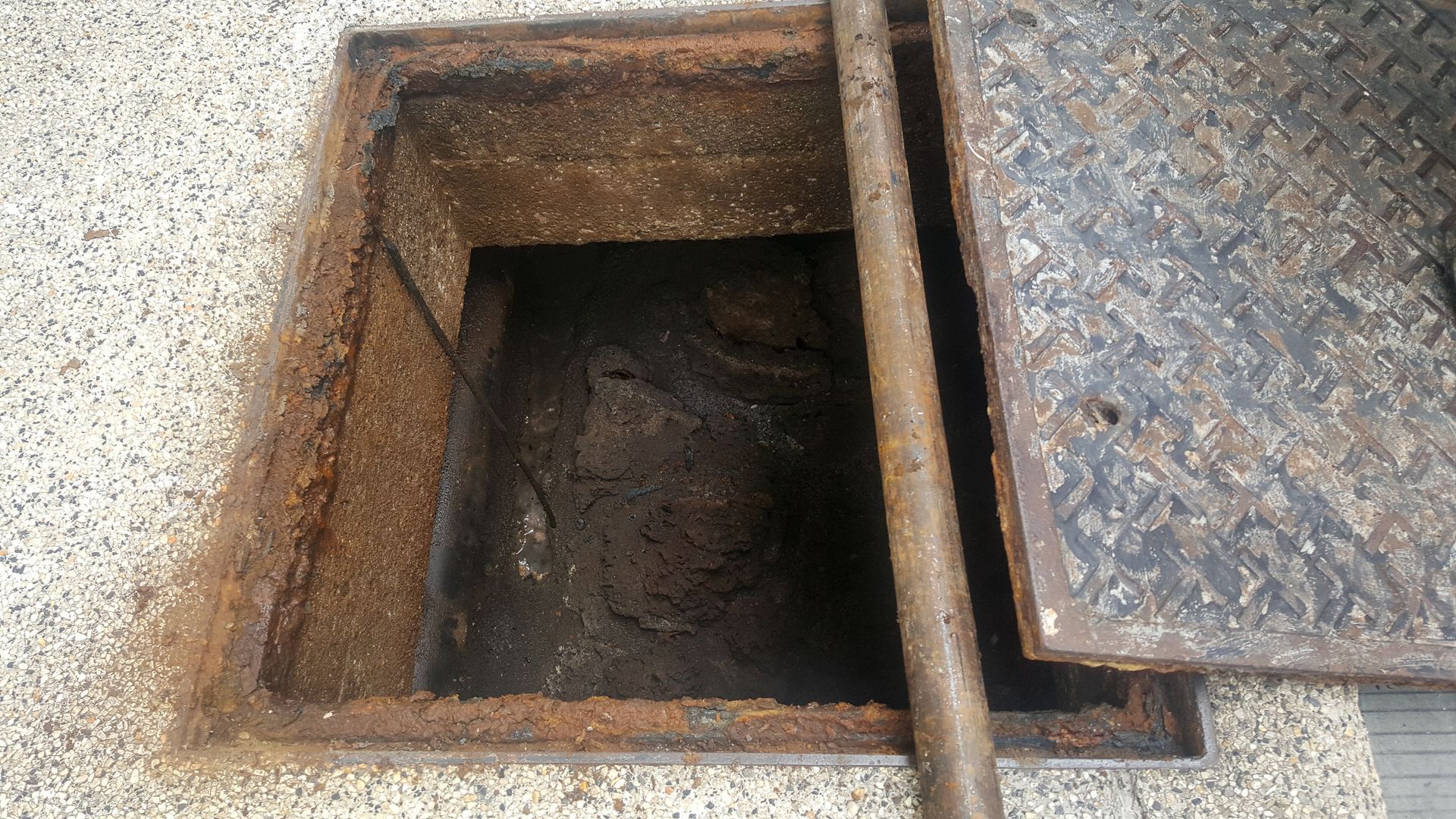 An open, square utility or drainage access hole in a concrete surface with a partially removed diamond-plate metal cover.