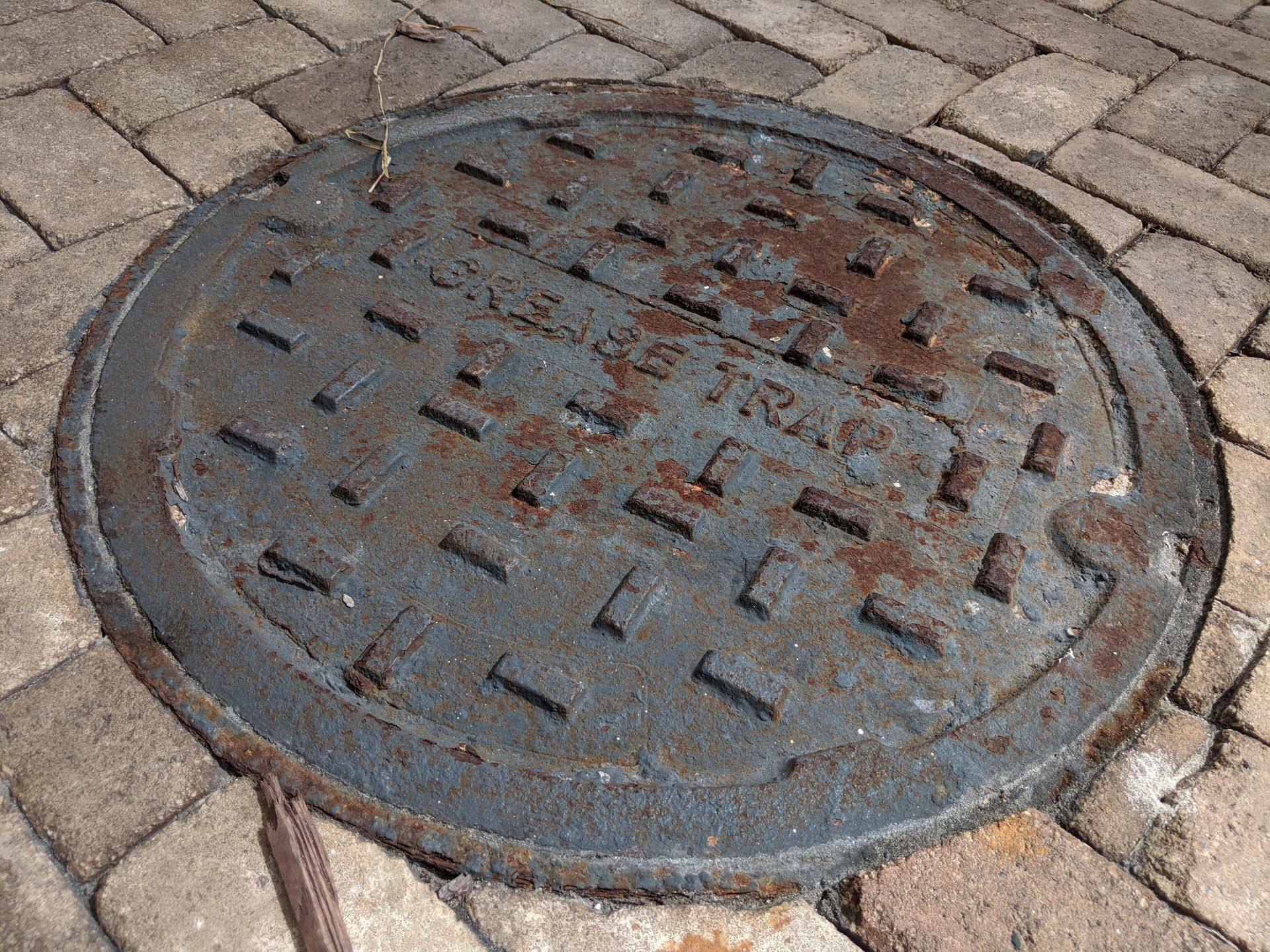 A round, rusted metal sewer cover with a grid pattern, set into a paved surface of rectangular stone blocks.