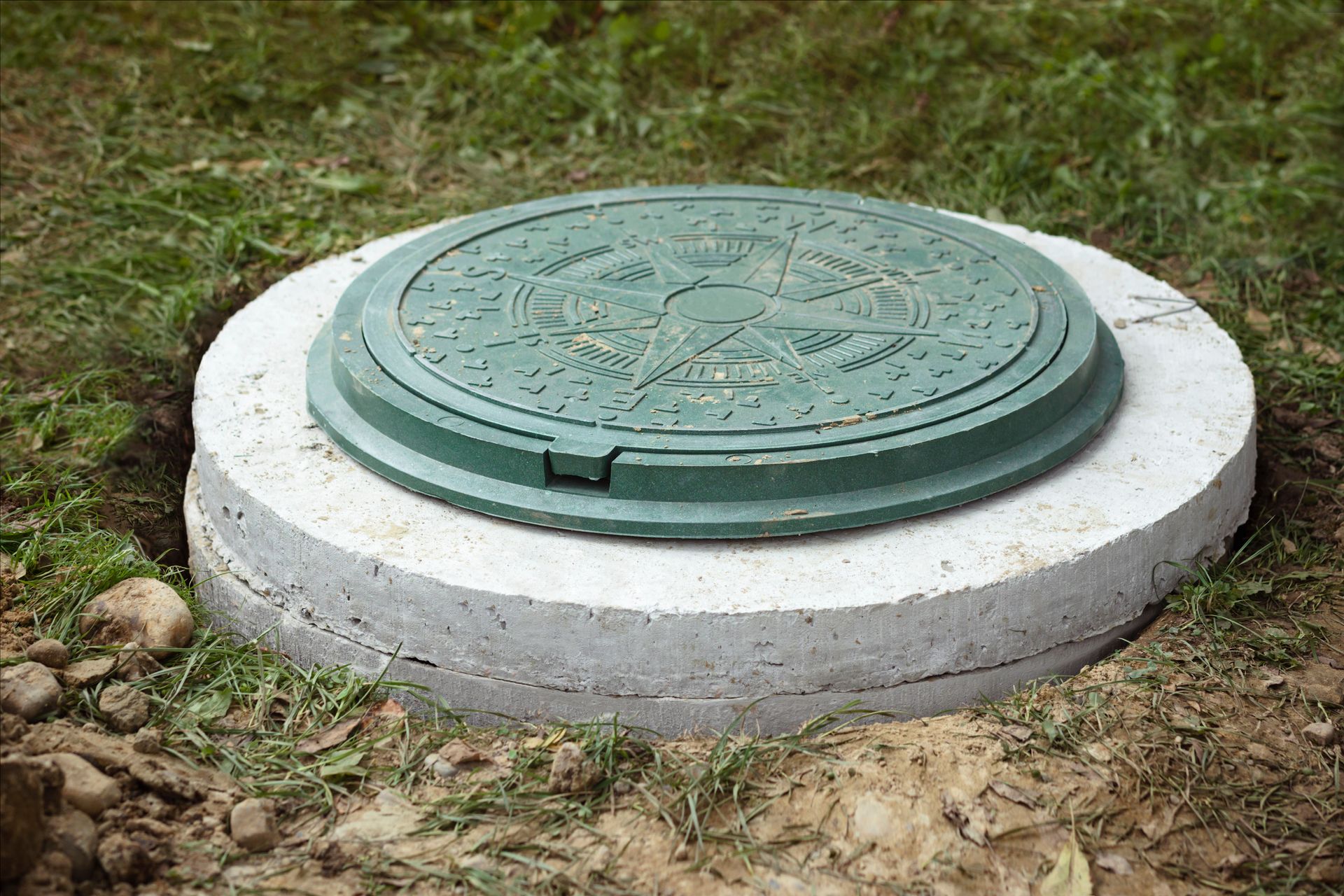 A green, circular manhole cover set in a white concrete ring, surrounded by dirt and grass.