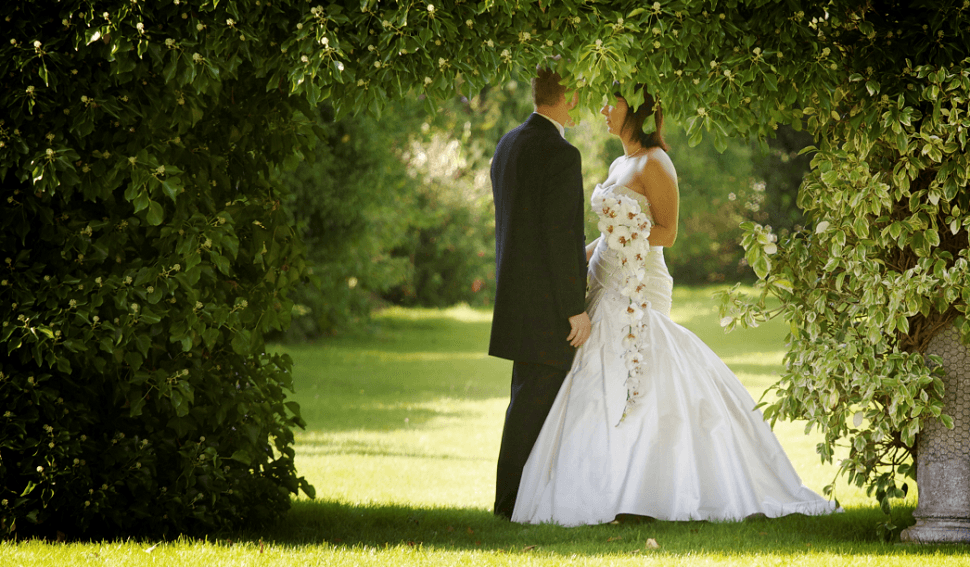A bride and groom under a garden arch; the bride is holding a lovely long train of flowers