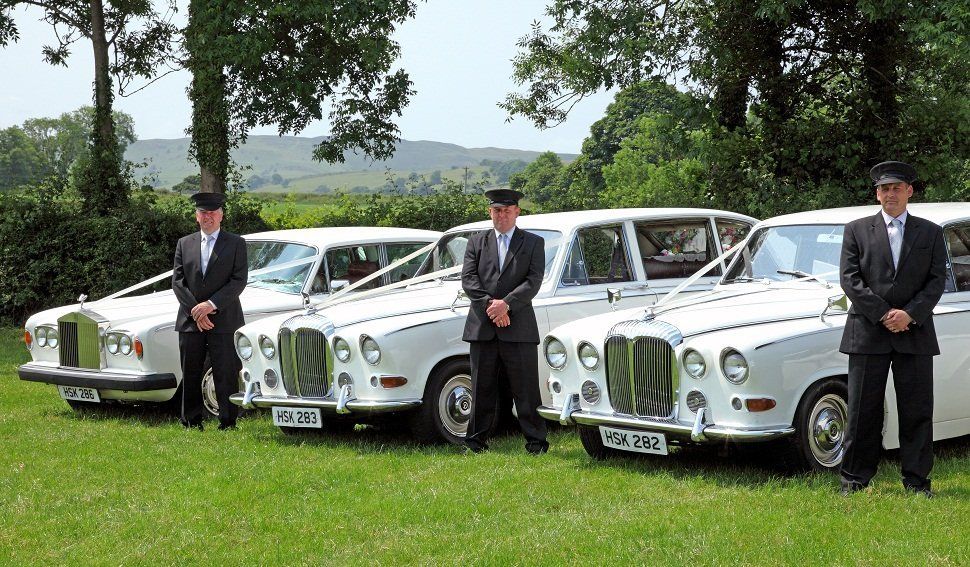 Three chauffeurs beside three white wedding cars