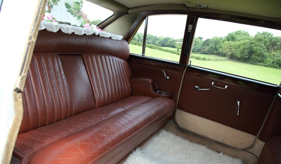 The back seat of a car furnished in red leather, with flowers in the rear window and a fluffy white carpet on the floor