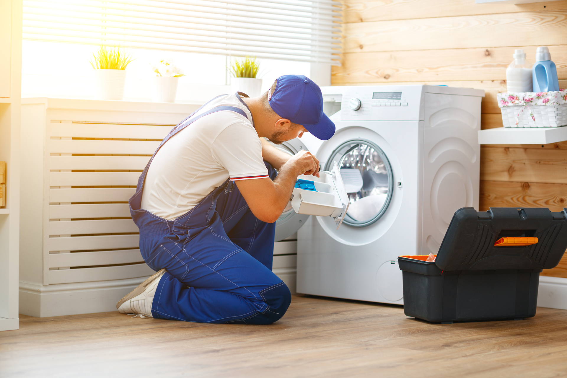 A repairman in blue overalls kneels, fixing a washing machine. A toolbox sits beside him in a laundry room.