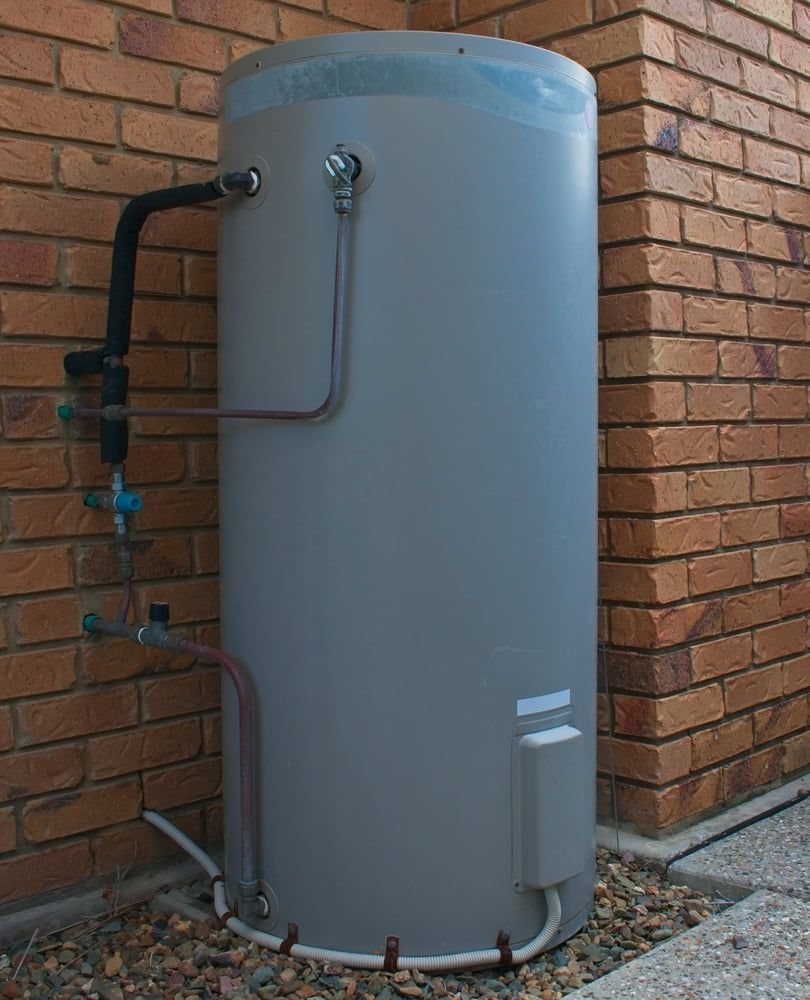 Grey Cylindrical Water Heater Beside a Brick Wall, With Exposed Pipes and Gravel — Mark's Plumbing in Noraville, NSW
