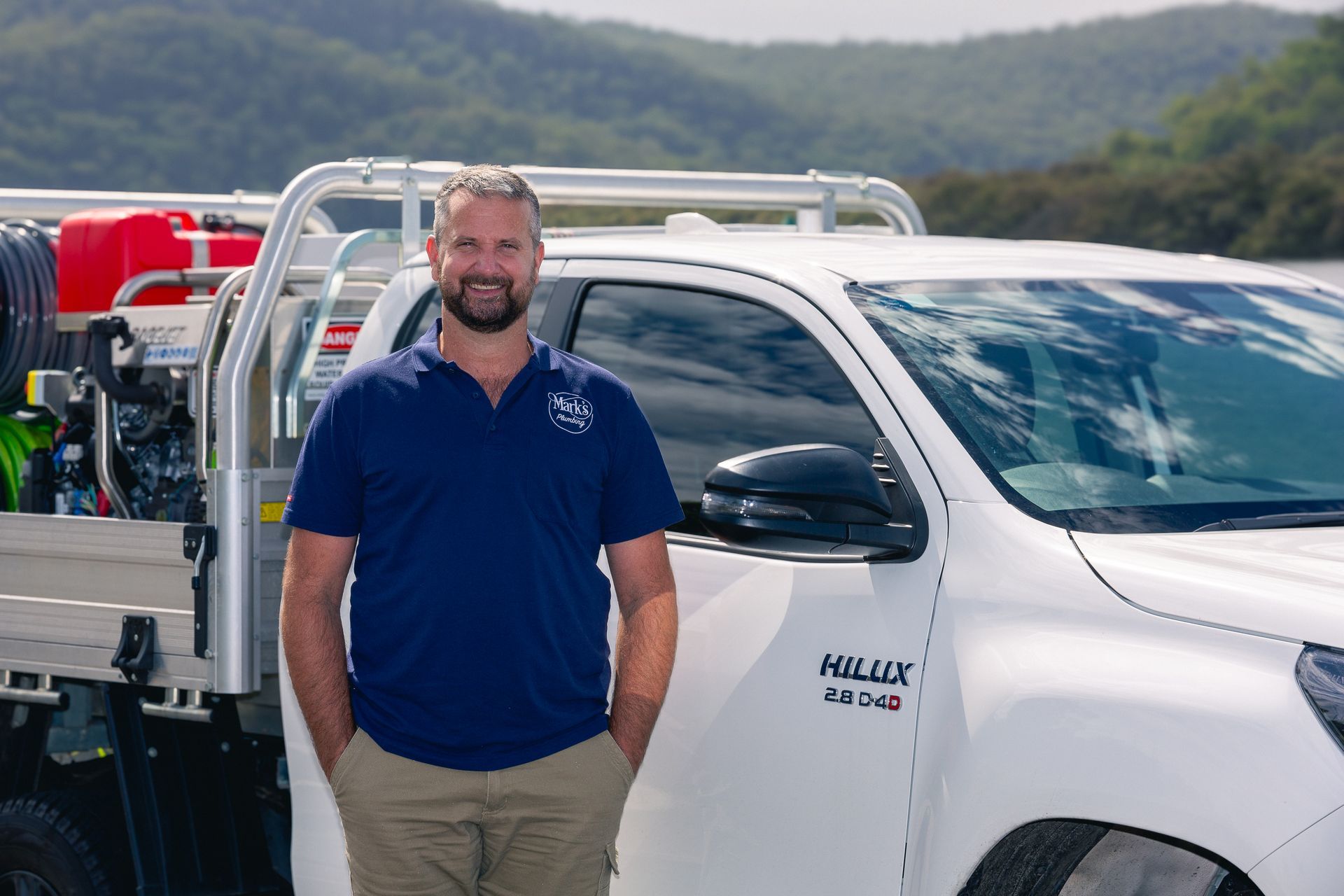Man Standing Beside a White Work Truck With a Flatbed, Outdoors in a Mountainous Area — Mark's Plumbing in Noraville, NSW