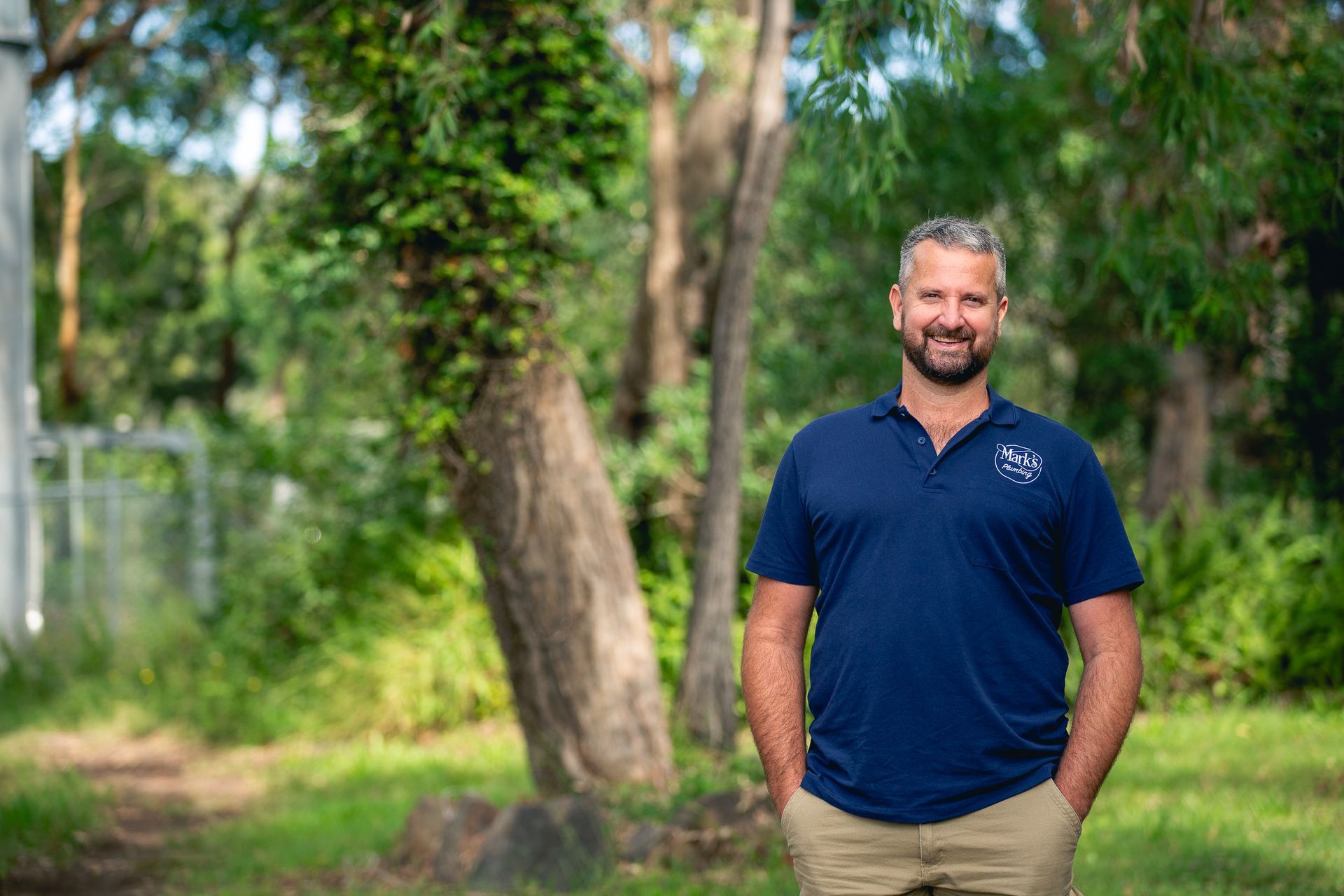 Man in a Navy Polo Shirt Standing Outdoors in a Green, Wooded Area, Smiling — Mark's Plumbing in Noraville, NSW