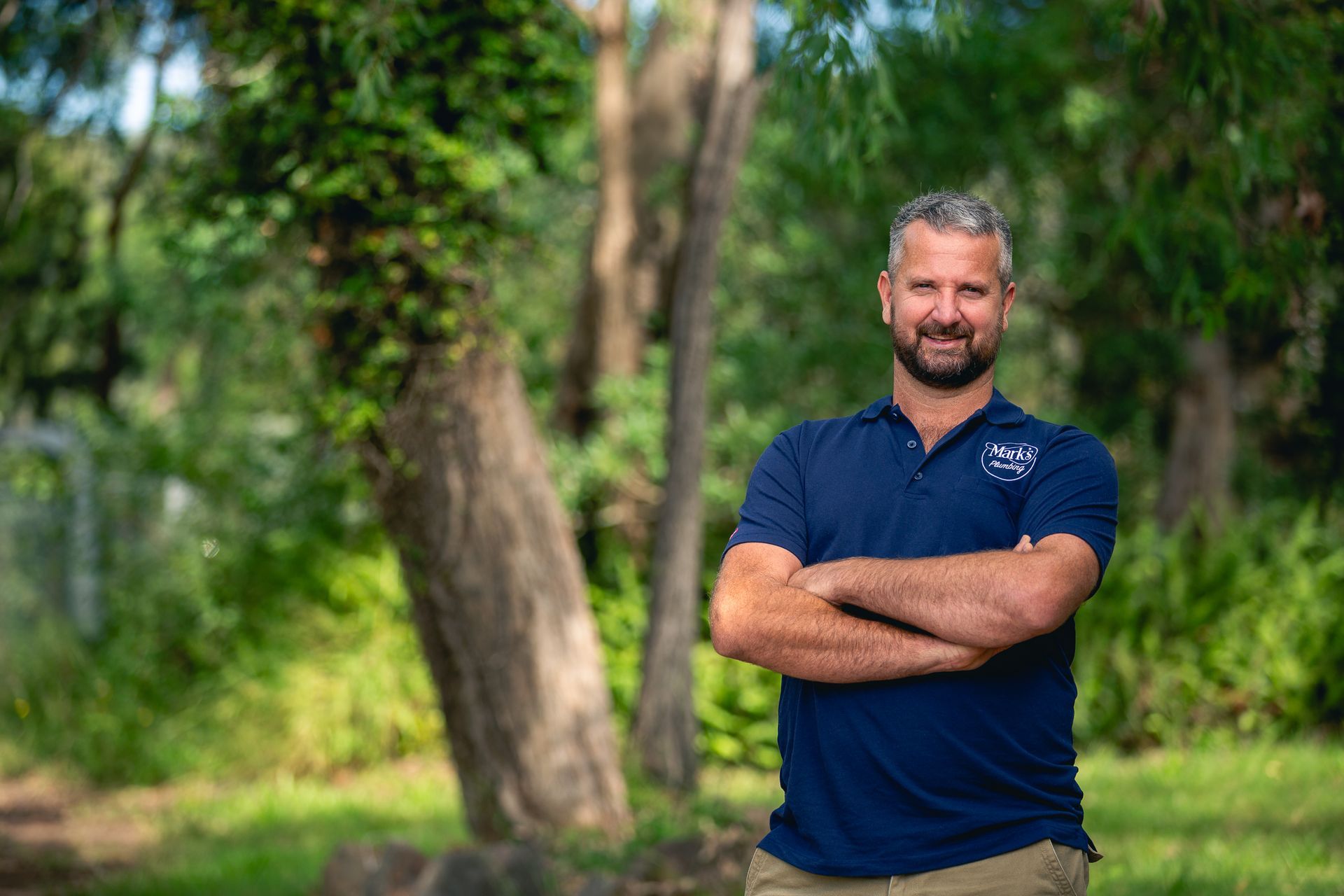 Smiling Man in a Navy Polo Stands With Arms Crossed in a Green Wooded Park — Mark's Plumbing in Noraville, NSW