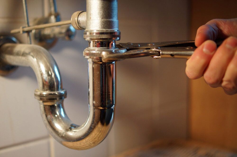 Hand Tightening a Wrench on a Shiny Chrome Sink Drain Pipe Under a Sink — Mark's Plumbing in Noraville, NSW