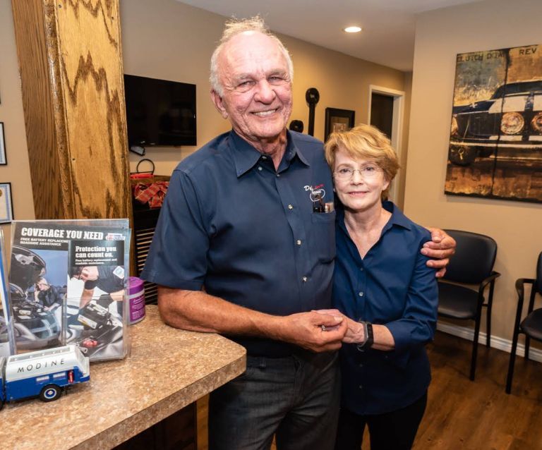 Two people smiling and posing together in a room beside a countertop display. | Dub's Garage