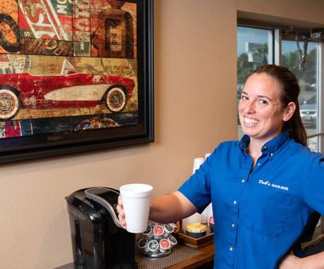 Woman in blue shirt holding a white cup beside a coffee station and framed car artwork. | Dub's Garage