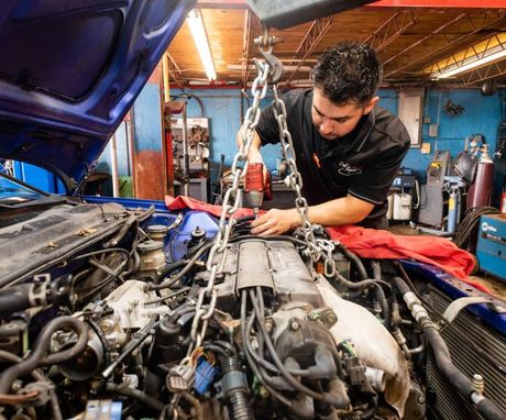 Mechanic working on car engine in a garage, leaning over the open hood with tools and chain hoist. | Dub's Garage