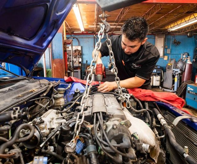 Mechanic working on car engine in a garage, leaning over the open hood with tools and chain hoist. | Dub's Garage