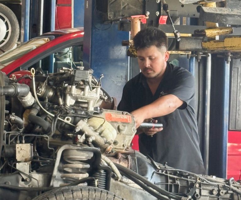 Mechanic working on a car engine in an auto repair shop with a red vehicle in the background | Dub's Garage