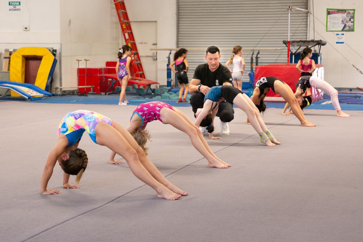 Gymnastics class, girls doing bridge pose with coach; gym interior.
