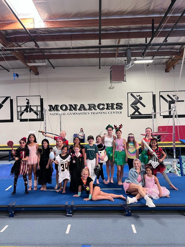 Group of costumed children and adults at a gymnastics studio, posing on a blue mat in front of a 