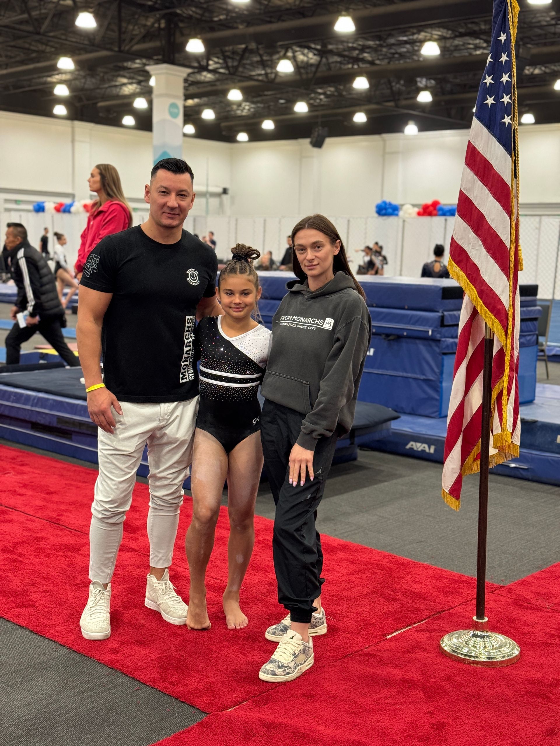 Man and two girls pose on red carpet near US flag, gymnastics equipment in background.