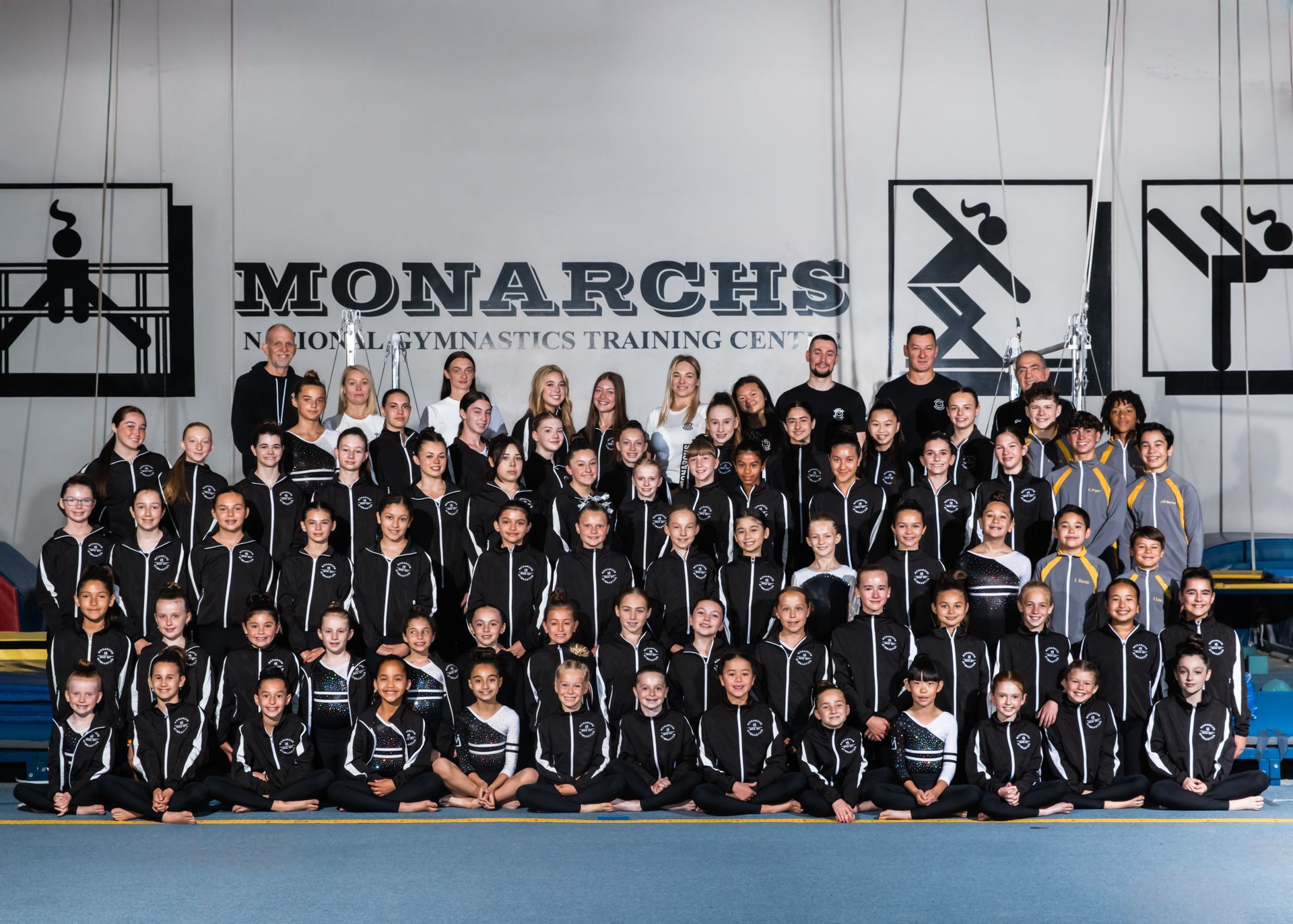 Group of gymnasts in matching black and white tracksuits posing in front of a gym banner that reads 