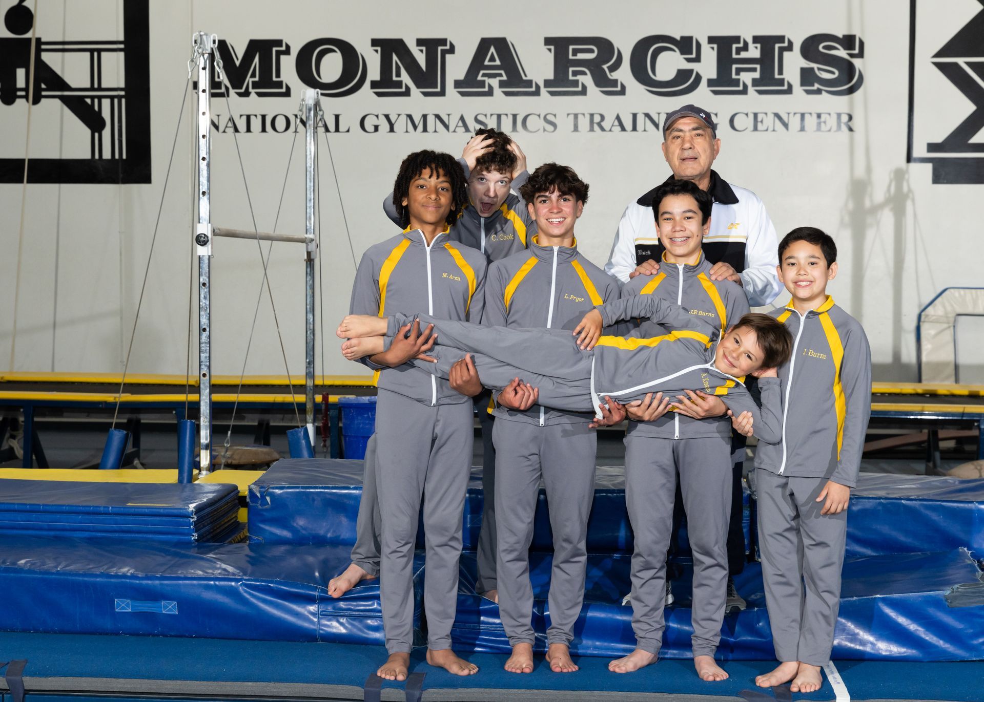 Gymnastics team in gray and yellow uniforms holding teammate. Team stands near equipment, Monarchs sign visible.