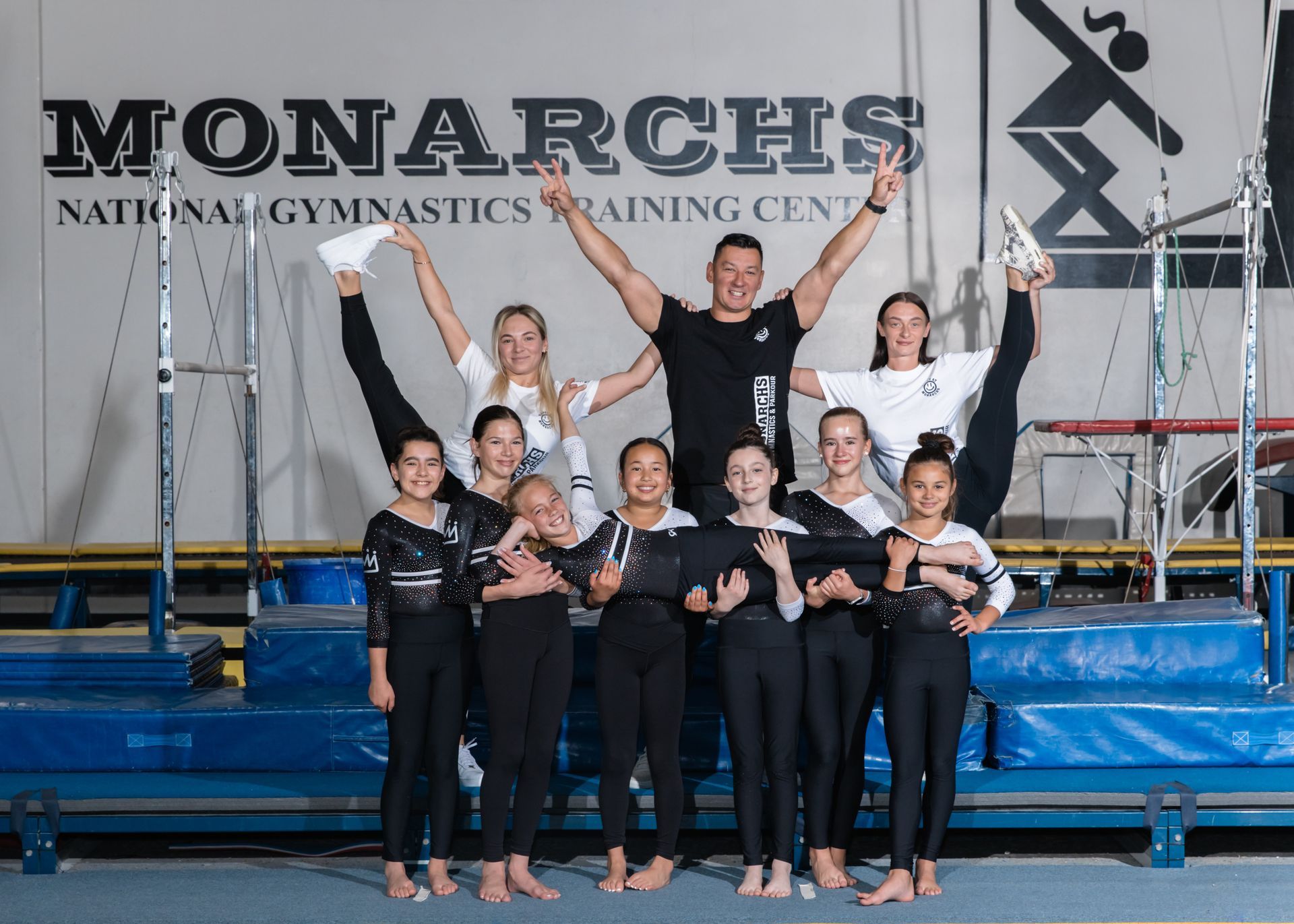 Gymnastics team poses at Monarchs National Gymnastics Training Center. Athletes in black leotards and coach pose with arms raised.