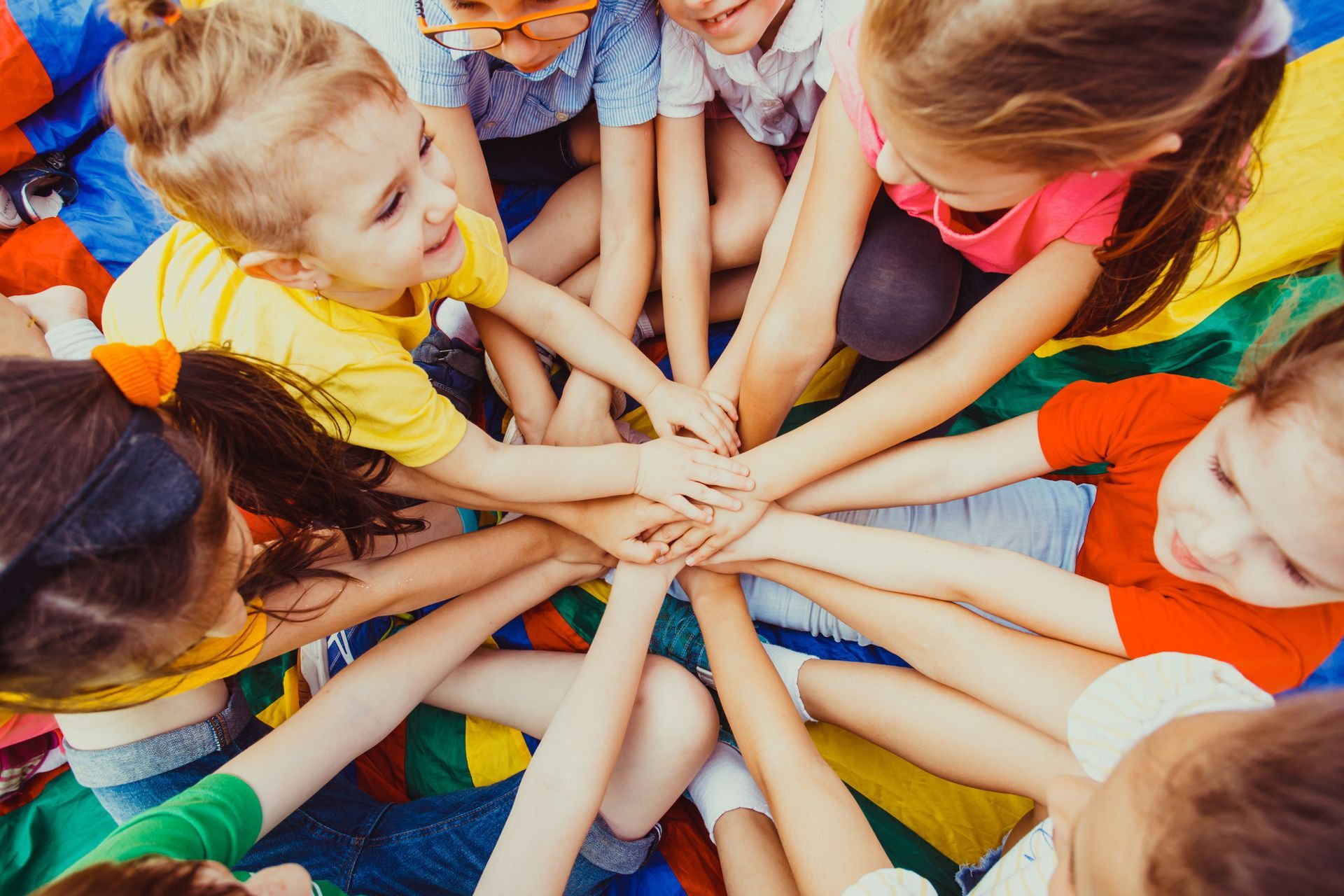 Children huddle together, hands in a circle. Smiling faces, colorful clothes, outside.