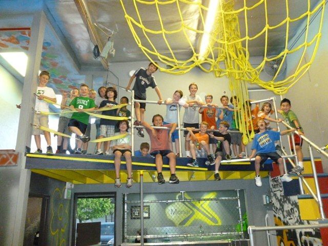 Children on a platform with yellow netting and bars; many look at the camera. Inside a building.