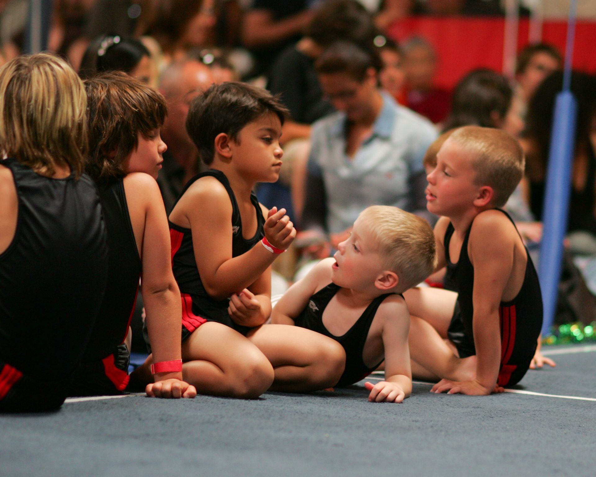 Gymnasts in black and red leotards on a blue mat, conversing. Spectators in the background.