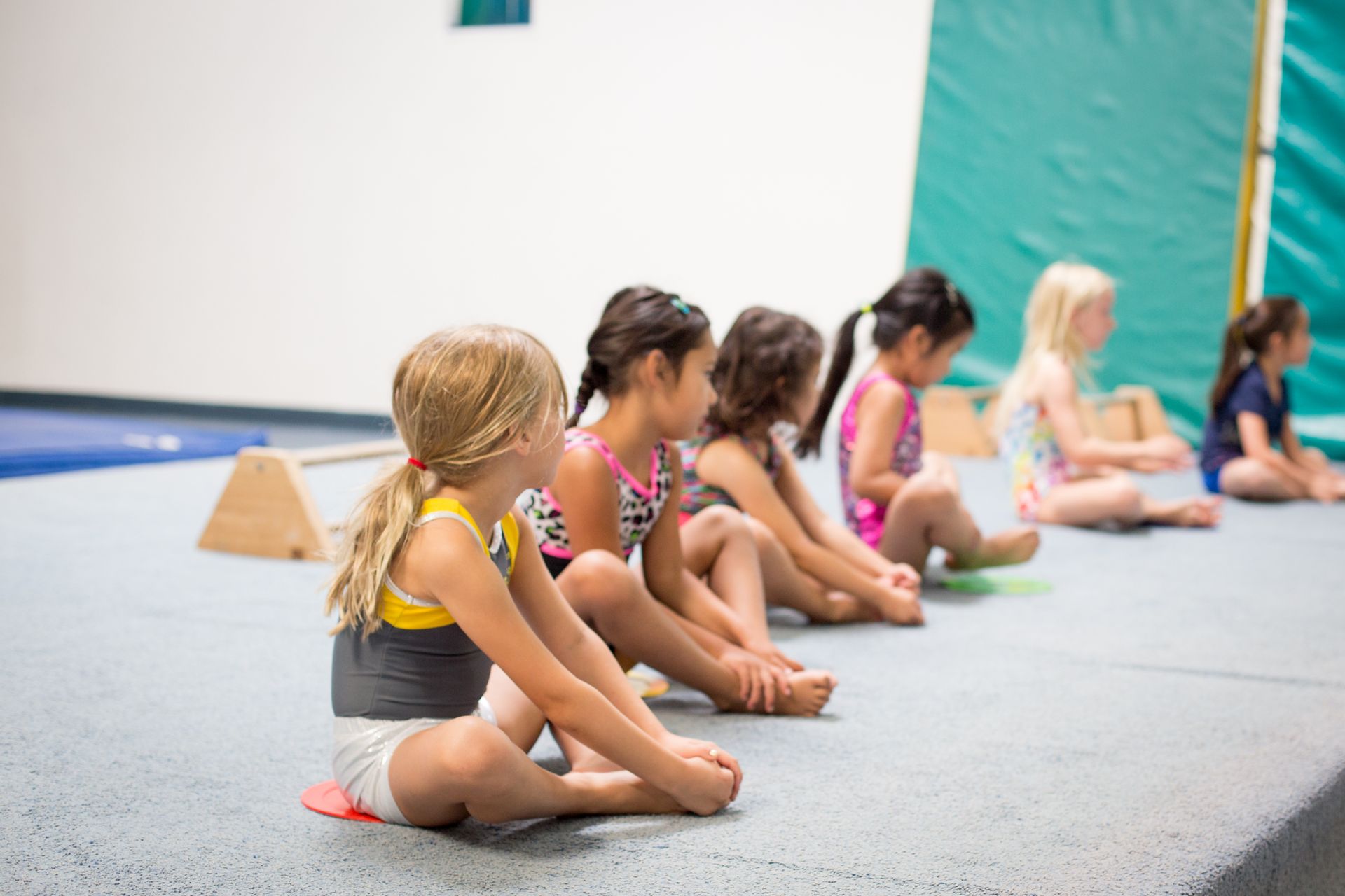 Girls in leotards sit on a gym floor, feet together, facing forward.