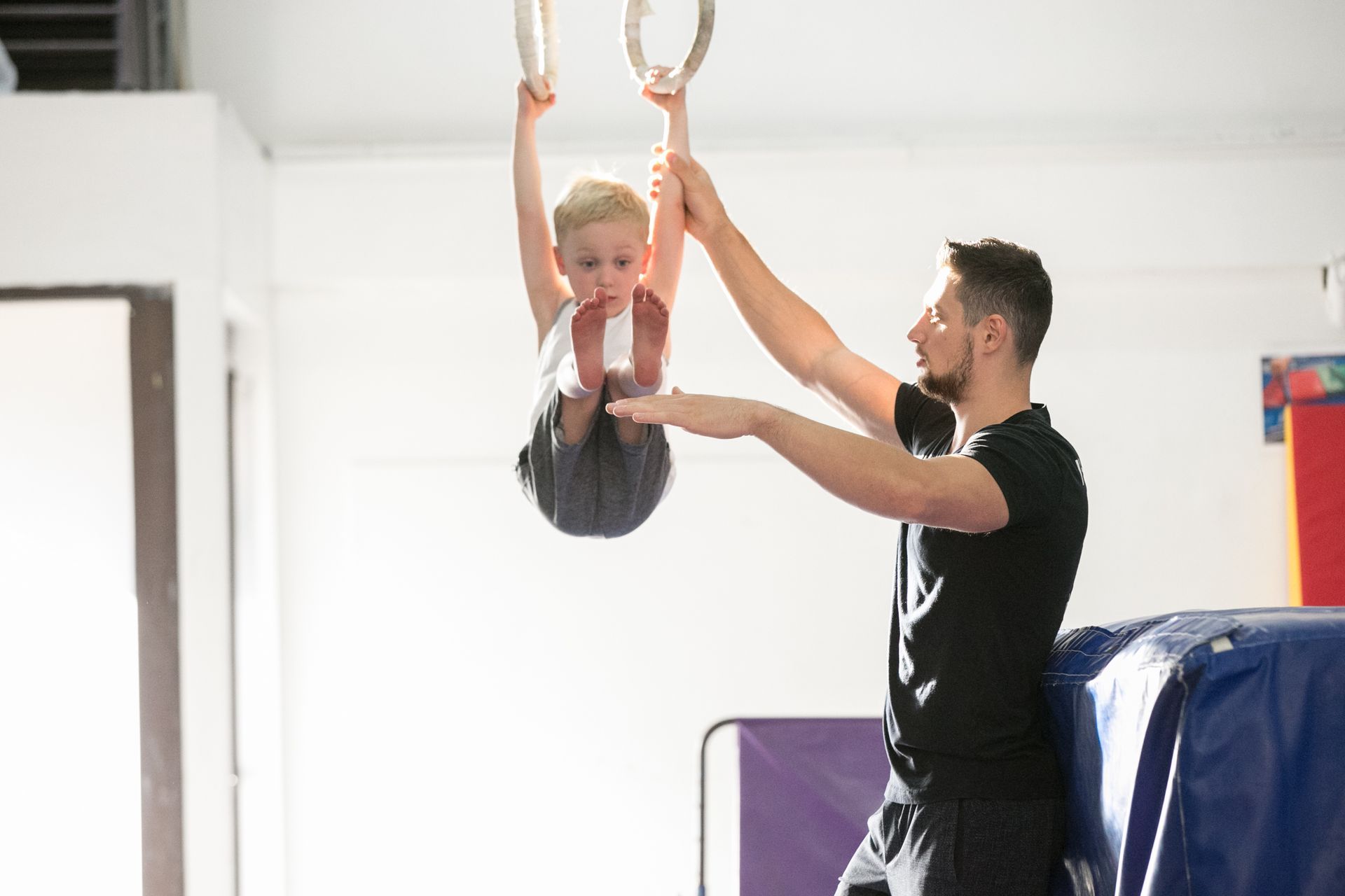 Boy hanging from gymnastic rings, instructor spotting him in a gym.