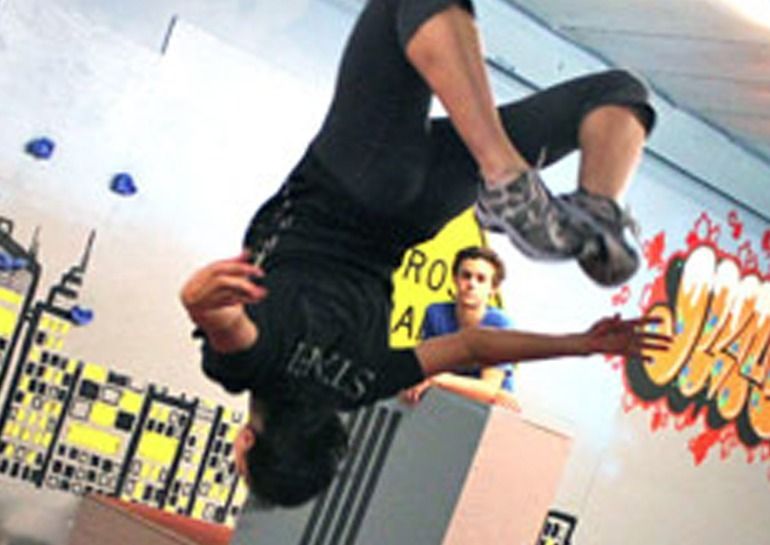 Person performing a backflip in an indoor parkour setting, with colorful graffiti and other people present.
