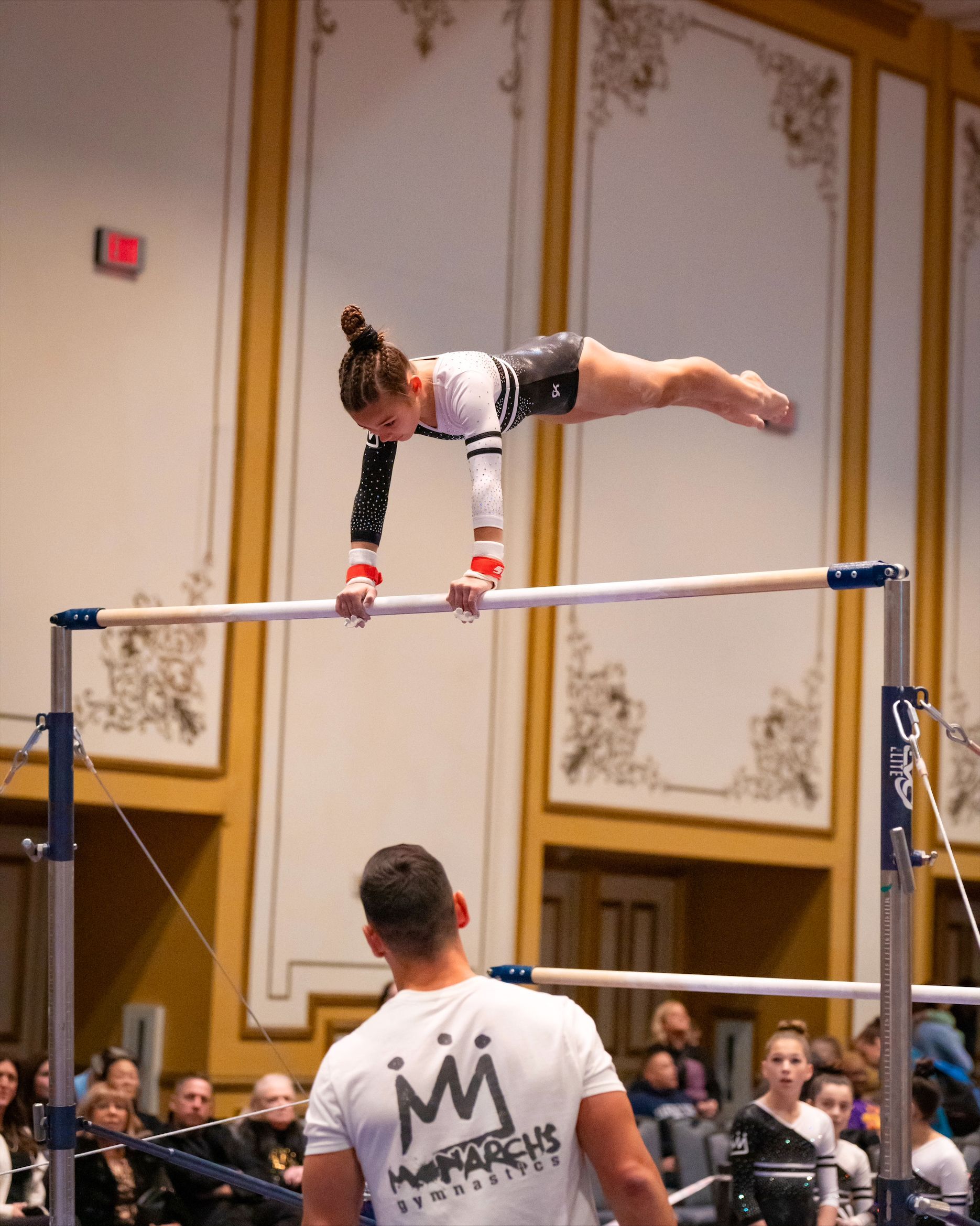 Gymnast on uneven bars performing a handstand, coach watches, gold-toned room.