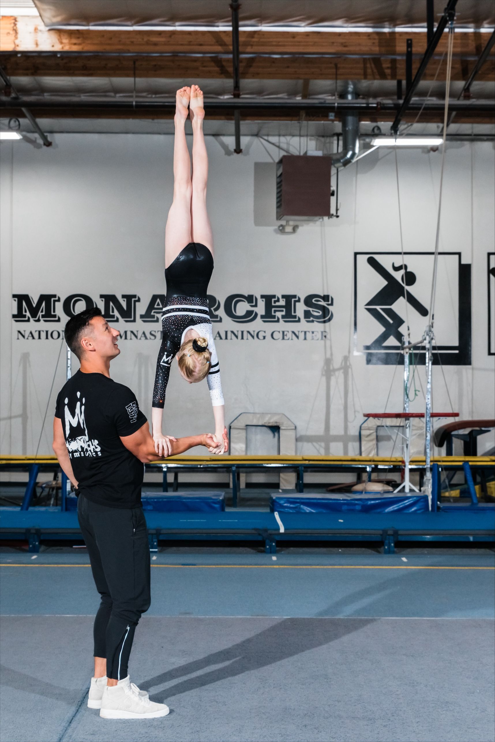 Gymnast doing handstand, supported by another person, inside a gym.