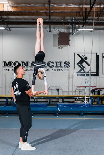 Gymnast doing handstand, supported by another person, inside a gym.
