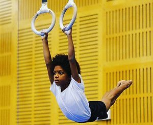 Young gymnast hanging from rings in a gymnasium, legs extended, wearing a white shirt and black shorts.