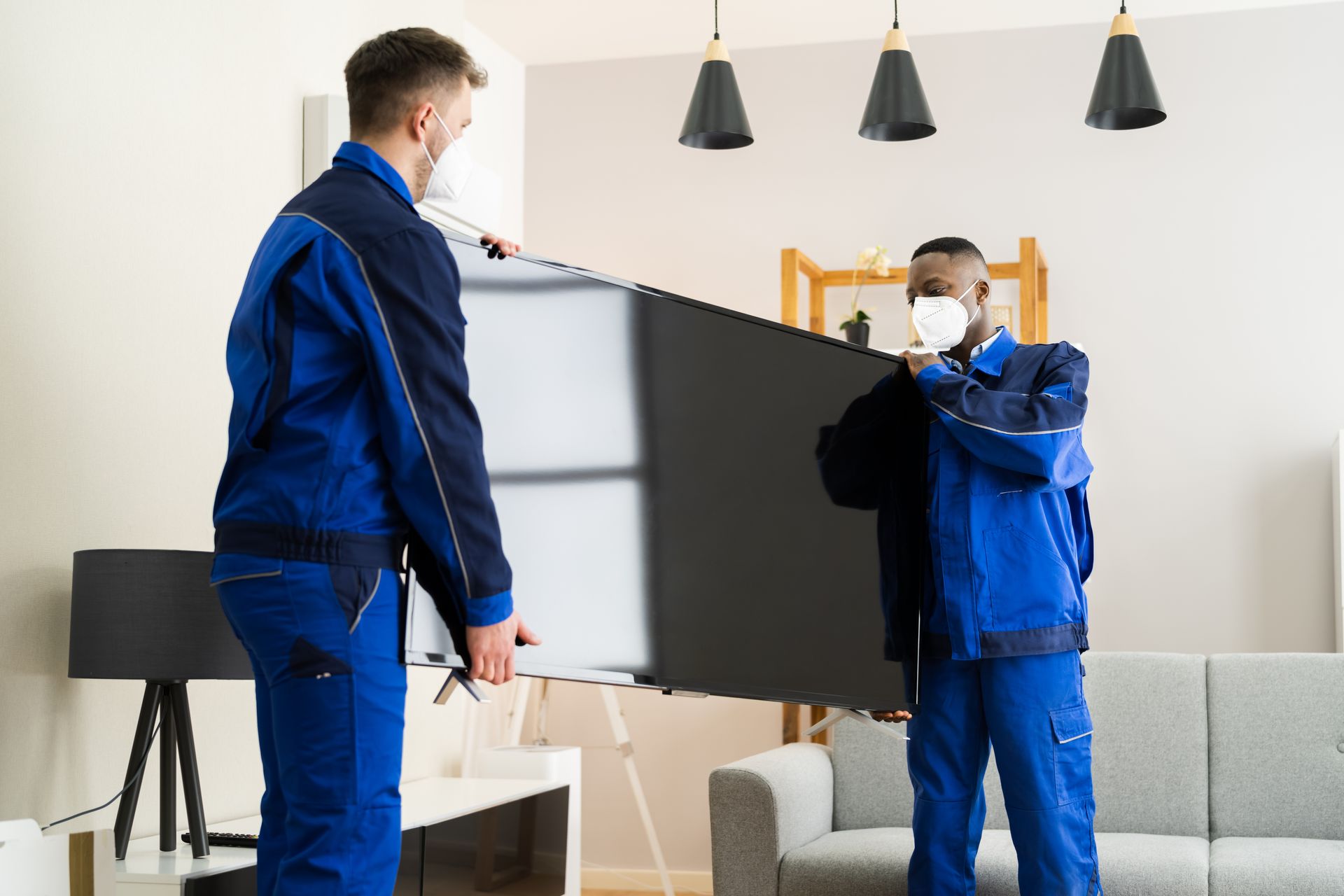 Two men wearing masks are carrying a flat screen tv in a living room.