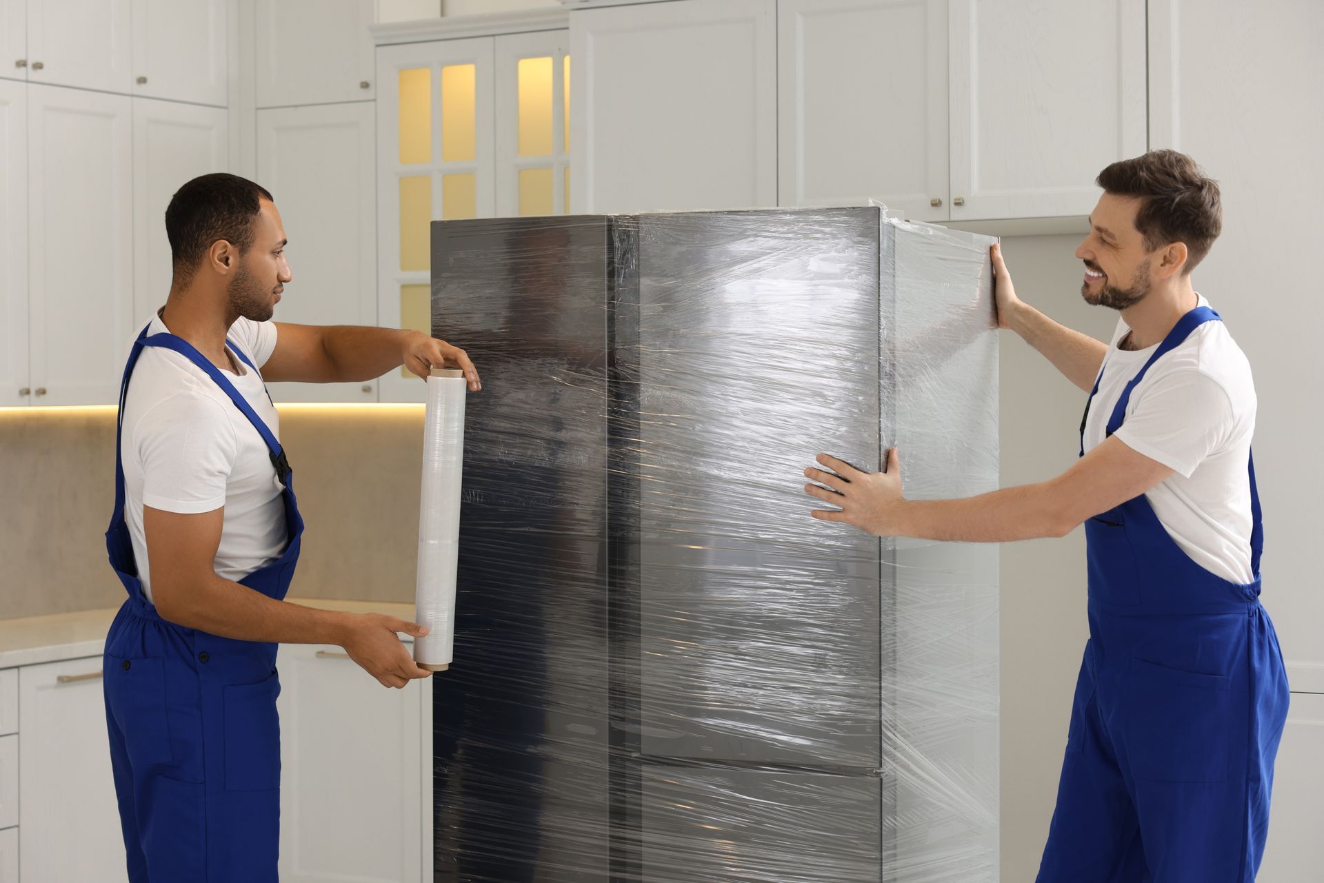 Two men are wrapping a refrigerator with plastic wrap in a kitchen.