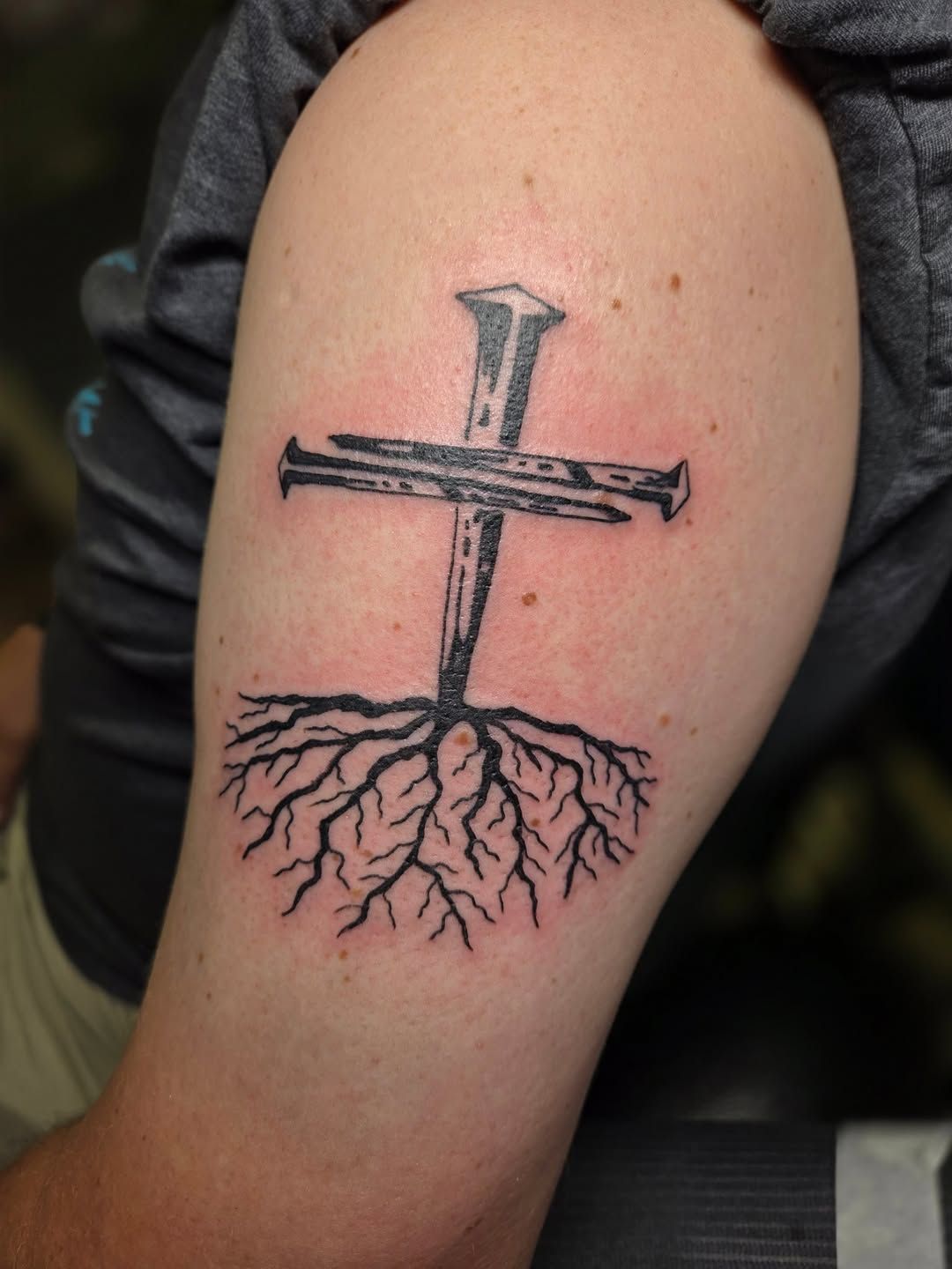 Tattoo of a cross made of nails with tree roots extending downward on a person's upper arm.