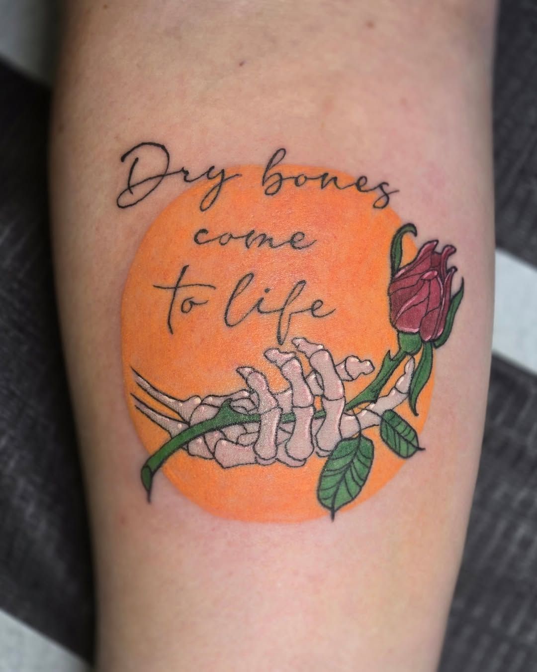 Tattoo: skeletal hand holding a rose, set against an orange circle, with the words 