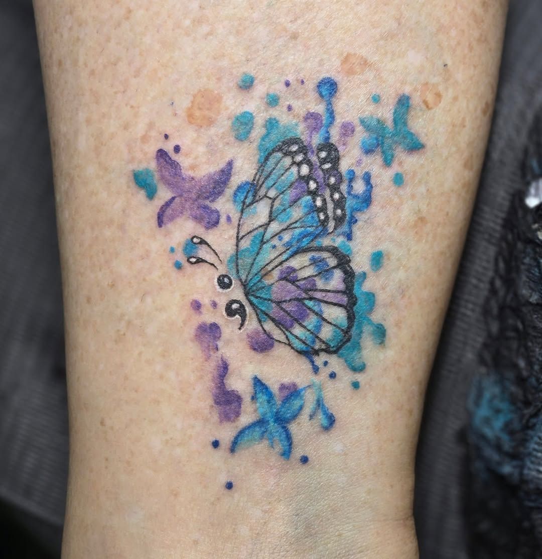 Butterfly tattoo on skin with watercolor-style blue and purple splashes, and semicolon.
