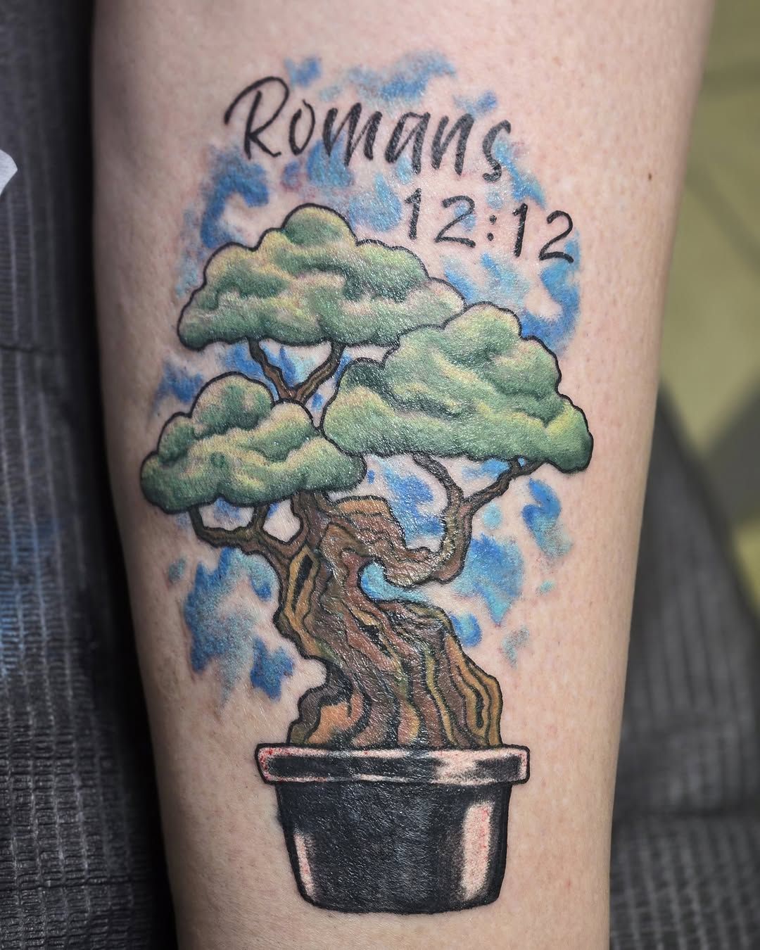 Tattoo of bonsai tree in a pot, with scripture Romans 12:12 in blue watercolor background on arm.