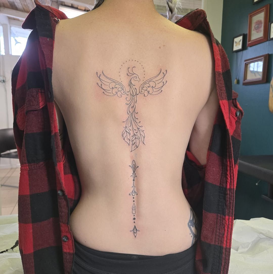Woman's back with a detailed spine tattoo. She's wearing an open red plaid shirt in a tattoo studio setting.