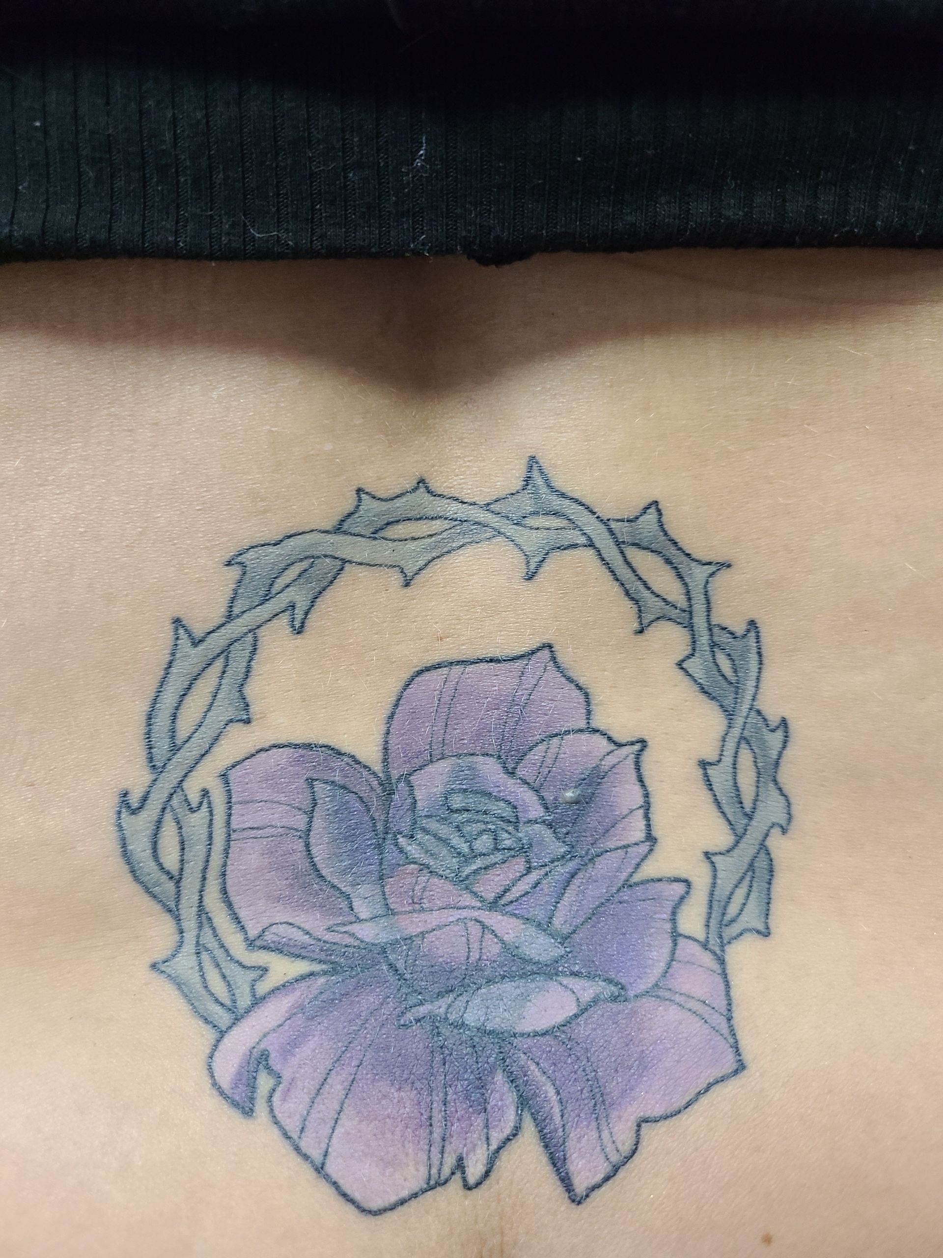 A person has a tattoo of a purple rose in a circle of thorns on their back.