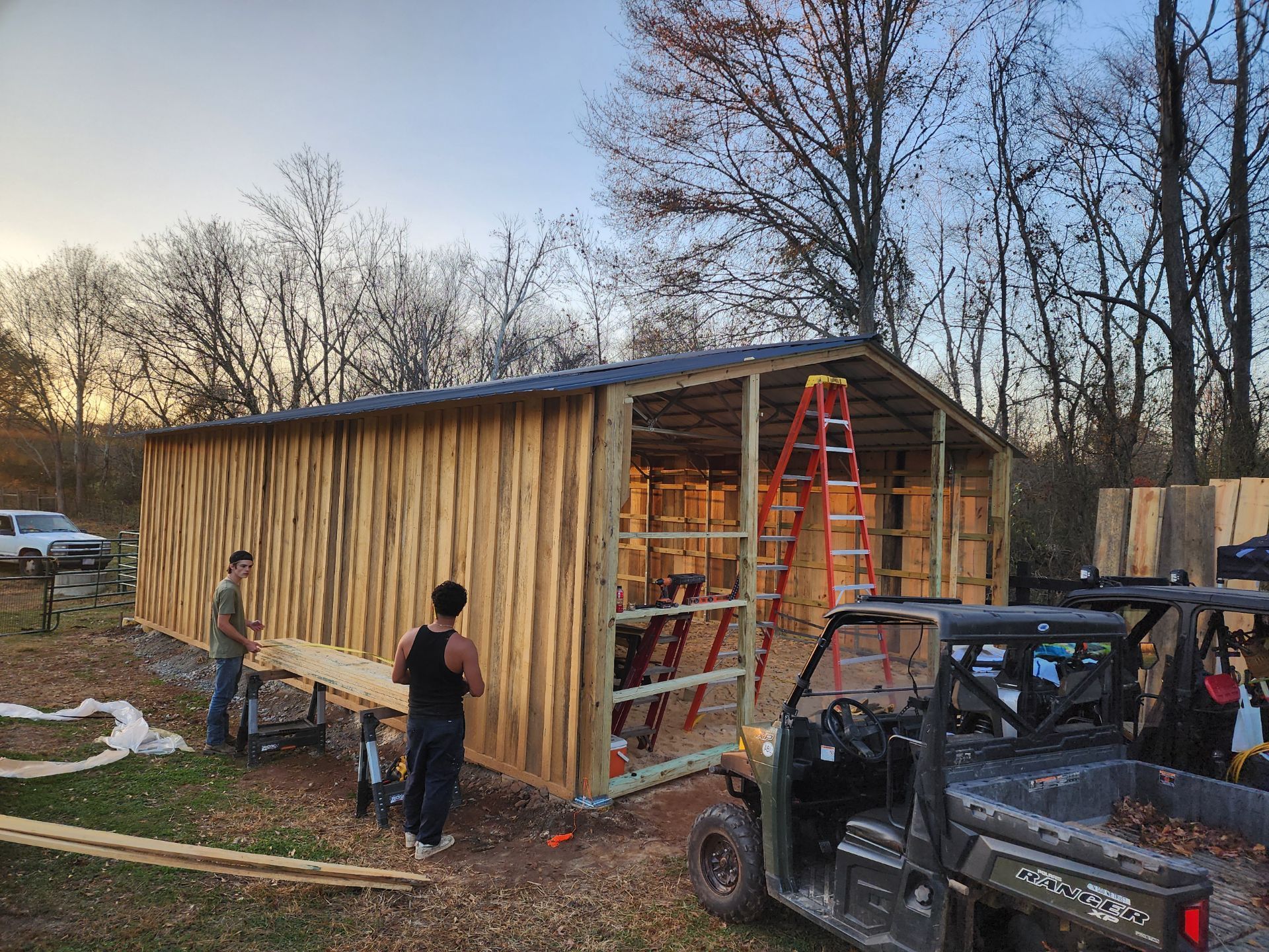 People constructing a wooden shed outdoors, with a utility vehicle nearby and a ladder.